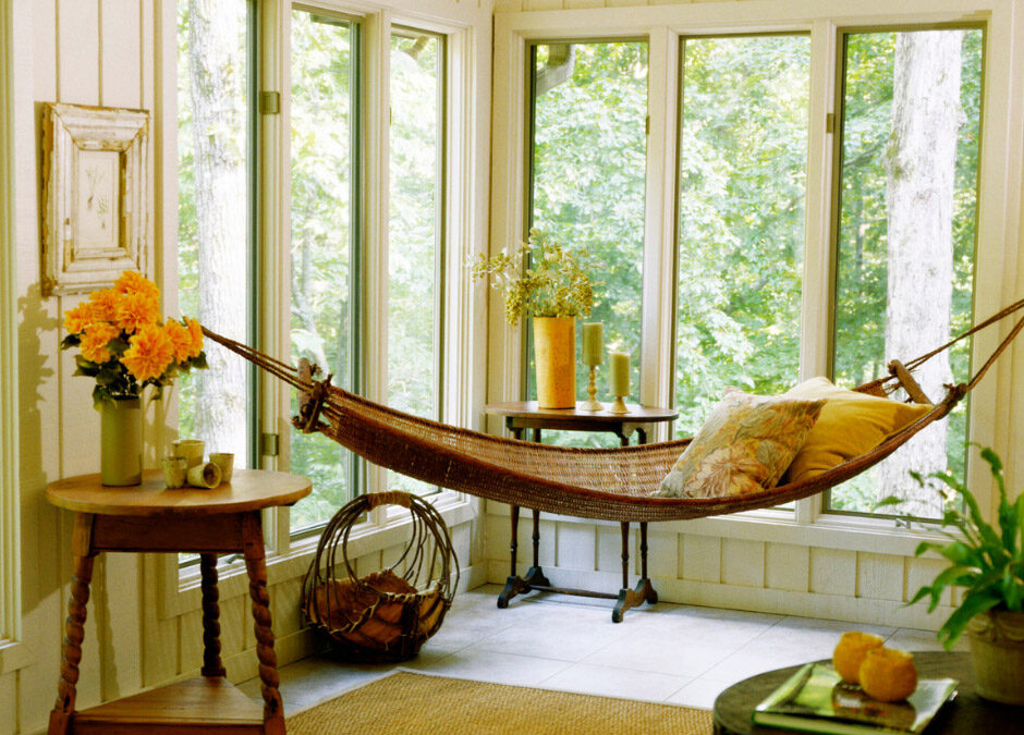 Bright sunroom with large windows, a woven hammock, wooden side tables, yellow flowers, and green trees outside