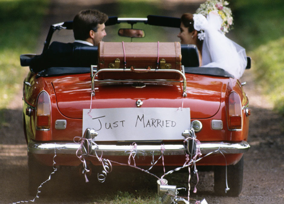 A newlywed bride and groom look at each other while driving away in a vintage red convertible down a dirt road. A brown suitcase is strapped to the luggage rack, and a handwritten "Just Married" sign with trailing tin cans decorates the back. - www.marquisandcoughlan.com